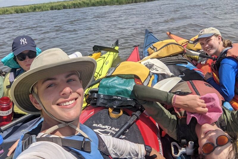 A group of people are enjoying a sunny day out on the water in their kayaks. They are smiling and appear to be having a good time. The water is calm, and the sky is clear. Everyone is wearing hats and life jackets, and they are all close together.